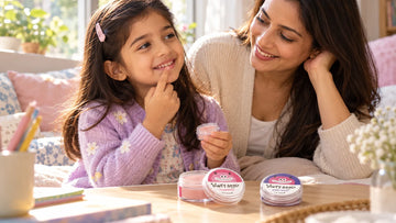 Girl dabbing Dirty Good Strawberry Kids Lip Balm from the tin with her fingertip while her mother watches, with a Berry Blast Kids Lip Balm tin on the table