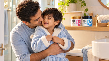 Father cuddling his son in a hooded bathrobe, with Dirty Good Pink Petal Kids Body Butter and Nautical Breeze Kids Body Wash on the shelf